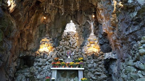 A stone grotto featuring a statue of a seated figure, flanked by two angels and adorned with flowers and candles.