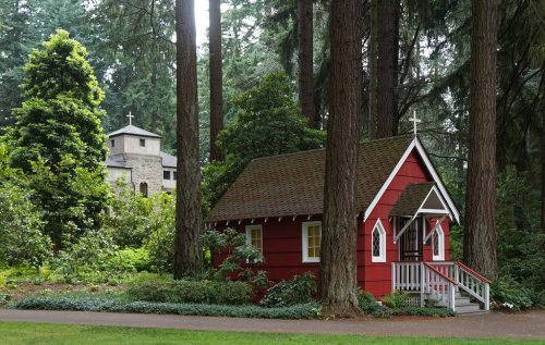 A small red chapel with a white porch surrounded by tall trees, with a larger stone building visible in the background.