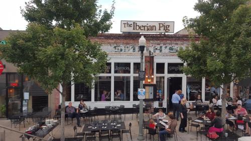 Outdoor dining scene at The Iberian Pig restaurant, with patrons seated at tables under trees on a lively street.