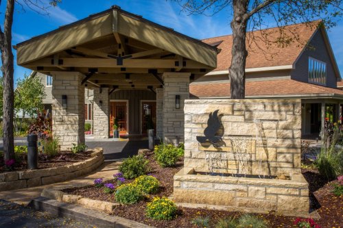 Entrance of a building with a stone facade, water feature, and colorful flowers under a clear blue sky.