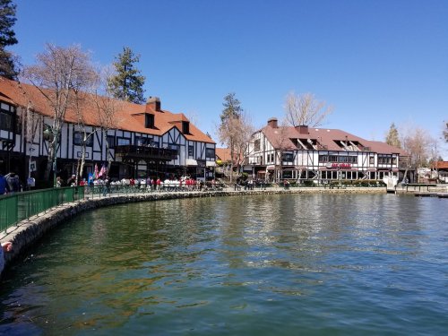 Scenic lakeside view featuring charming buildings and a clear blue sky. Trees line the shore, reflecting in the water.