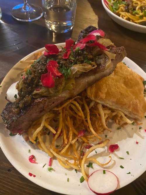 A plate featuring a grilled steak topped with herbs and flowers, served with crispy fries and a flaky pastry.