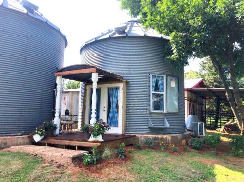Two metal silos converted into cozy homes, featuring a porch with white columns and flower pots, surrounded by greenery.