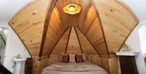 Cozy bedroom with a wooden, sloped ceiling, featuring a bed with decorative pillows and a warm light fixture.