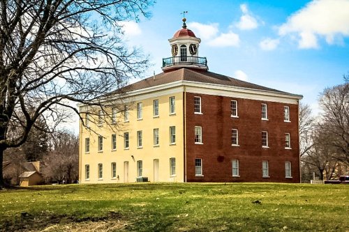Historic building with a red and yellow facade, featuring a cupola, surrounded by green grass and trees under a blue sky.