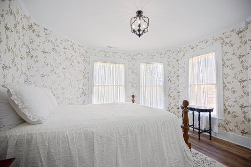 Cozy bedroom with a white bed, floral wallpaper, and large windows letting in natural light.