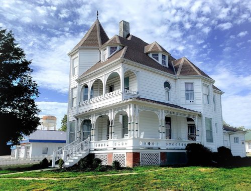 A large, white Victorian-style house with a turret, surrounded by green grass and a blue sky with clouds.