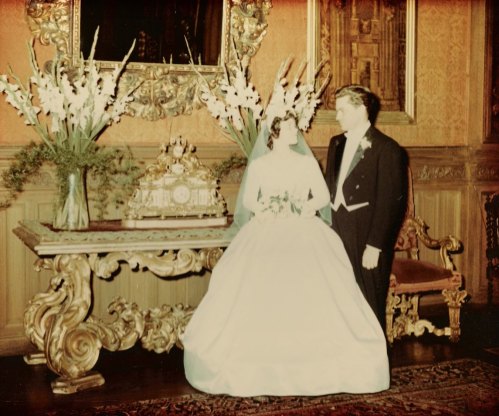 A bride in a white gown and a groom in a tuxedo stand together in an ornate room with floral arrangements.