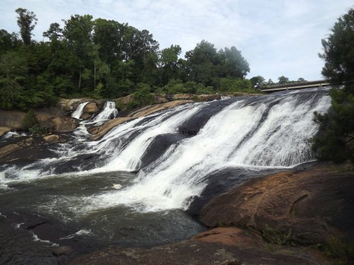 A cascading waterfall flows over rocky terrain, surrounded by lush green trees under a cloudy sky.