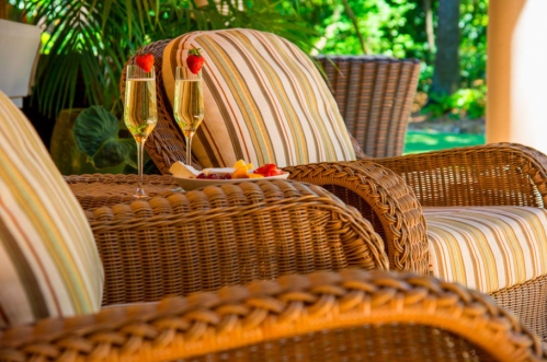 Two glasses of champagne with strawberries sit on a table beside wicker chairs, surrounded by greenery.