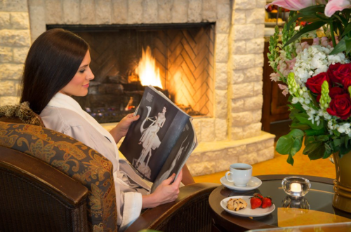 A woman in a robe sits by a fireplace, reading a magazine with a cup of coffee and a plate of cookies and strawberries nearby.
