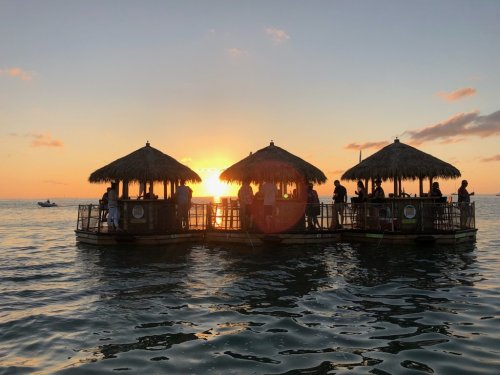 Silhouetted tiki huts on water at sunset, with people enjoying the view and a boat in the distance.