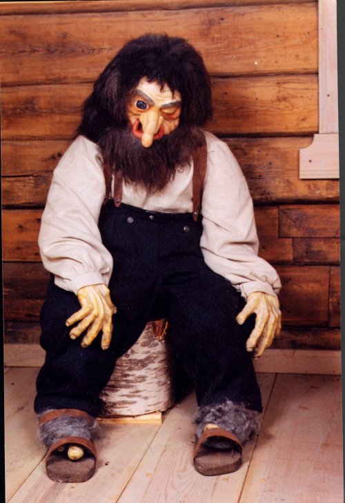 A lifelike troll figure with a large nose and beard, sitting on a wooden stool in a rustic setting.