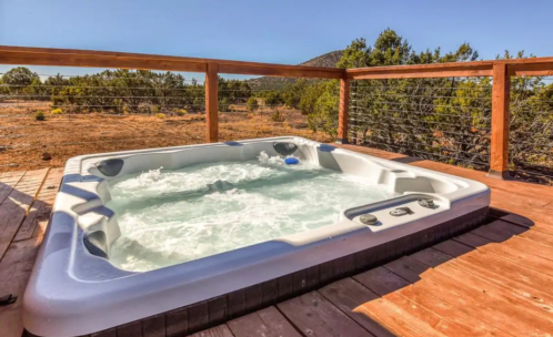 A hot tub on a wooden deck overlooking a dry landscape with trees and mountains in the background.