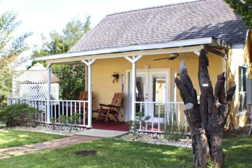 A cozy porch with rocking chairs, surrounded by greenery and a tree, in front of a charming yellow house.