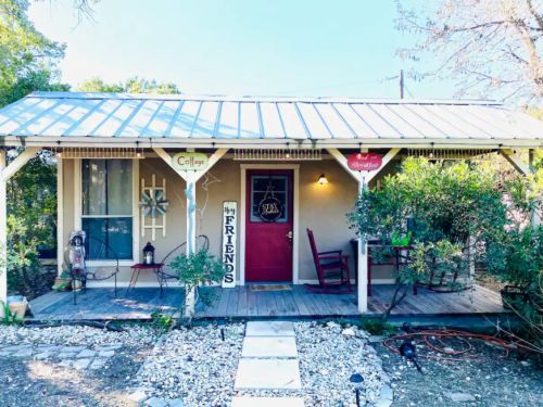 Charming cottage with a red door, porch, and signs welcoming friends, surrounded by greenery and gravel path.