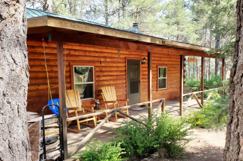 A cozy log cabin surrounded by trees, featuring a porch with two chairs and a lantern.