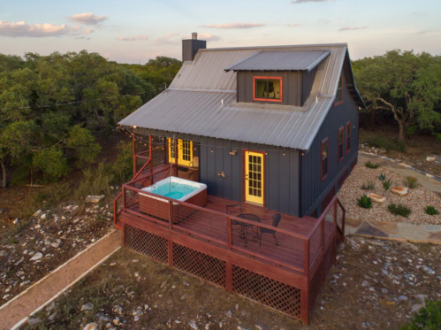 A cozy black cabin with a hot tub on a wooden deck, surrounded by trees and rocky terrain at sunset.