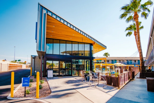 Modern building entrance with large glass windows, wooden accents, and palm trees under a clear blue sky.