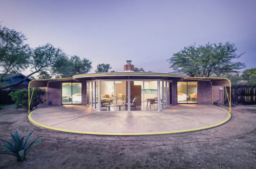 A modern round house with large glass windows, surrounded by desert landscaping at dusk.