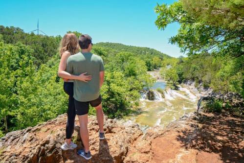 A couple stands together on a rocky ledge, overlooking a river and waterfall surrounded by lush greenery.