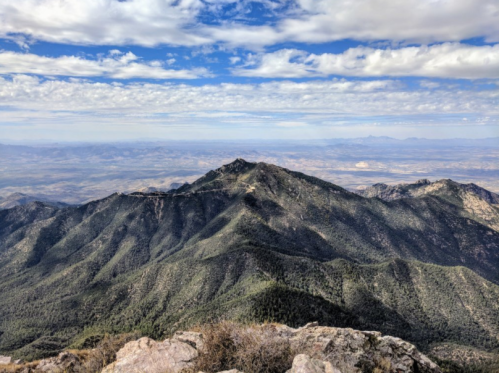 A panoramic view of rugged mountains under a blue sky with scattered clouds, showcasing lush green slopes and distant valleys.