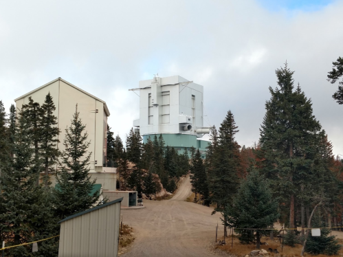 A tall white building surrounded by trees, with a dirt road leading up to it, under a cloudy sky.