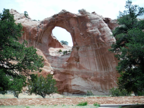 A large rock formation with a circular arch, surrounded by green trees and a stone wall.