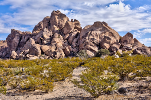 A rocky formation rises above a desert landscape, surrounded by sparse vegetation and a blue sky with scattered clouds.