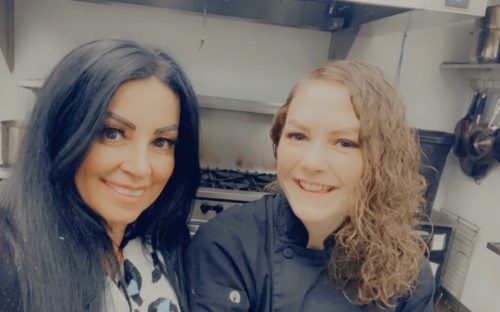 Two women smiling in a kitchen, one wearing a chef's coat, with cooking equipment in the background.