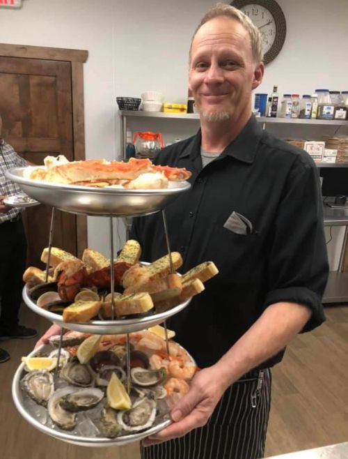 A smiling man in a black shirt holds a three-tiered seafood platter with shrimp, oysters, and crab legs.