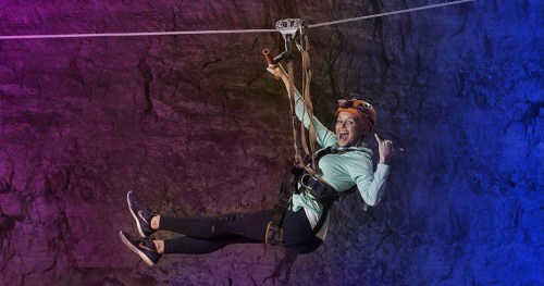 A person zip-lining with excitement, wearing a helmet and harness, against a colorful rocky background.