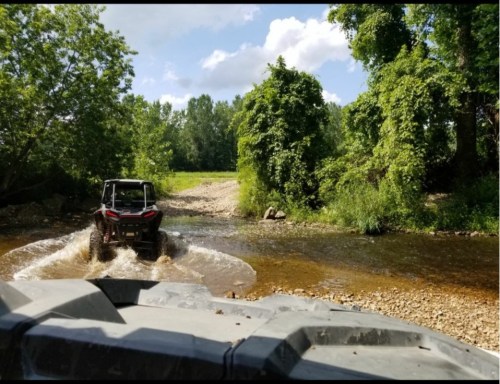 A vehicle splashes through a shallow creek surrounded by lush greenery and trees under a partly cloudy sky.