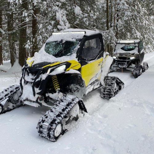 A yellow snowmobile parked in a snowy forest, with another snowmobile visible behind it, both covered in snow.