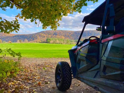 A side view of an ATV parked on a dirt path, with a scenic landscape of green fields and autumn-colored hills in the background.