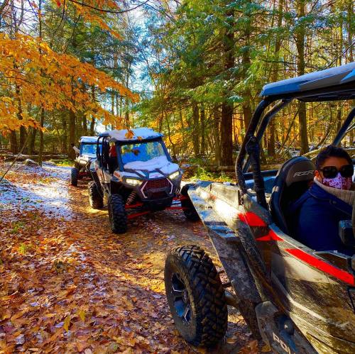 Two off-road vehicles on a forest trail surrounded by autumn leaves and trees, with a hint of snow on the ground.