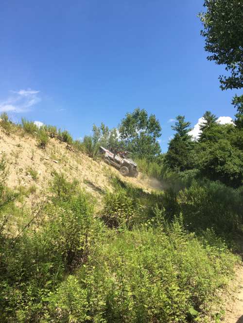 A vehicle climbs a sandy hill surrounded by greenery under a clear blue sky. Dust is kicked up from the tires.
