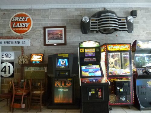A retro arcade area featuring vintage games, a classic car front, and nostalgic decor on a brick wall.