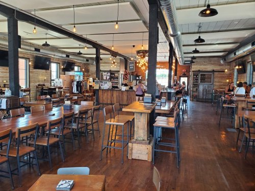A spacious, rustic restaurant interior with wooden tables, a bar area, and warm lighting. Patrons are seated and ordering.