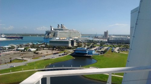 A view of a large cruise ship docked near a harbor, with green lawns and a parking lot in the foreground.
