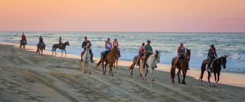 A group of people riding horses along a sandy beach at sunset, with gentle waves in the background.