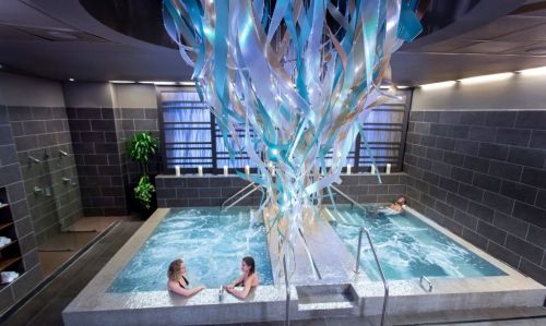 Two women relax in a spa with a large, colorful ribbon installation hanging above a bubbling hot tub.