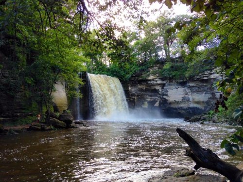 A serene waterfall cascades into a calm river, surrounded by lush green trees and rocky terrain.