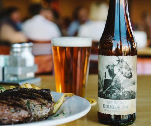 A bottle of Hop Rising Double IPA next to a glass of beer and a plate of steak with fries in a restaurant setting.