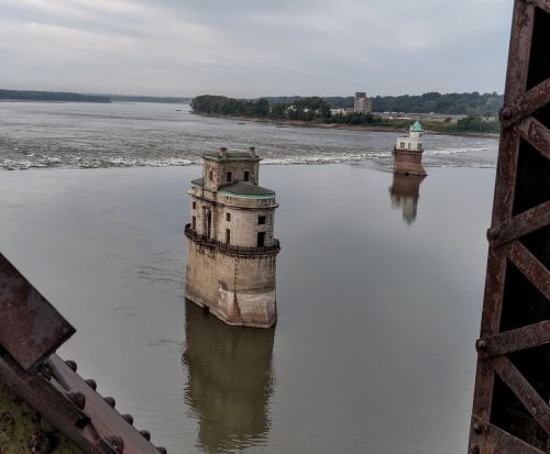 Two stone towers rise from calm water, surrounded by greenery under a cloudy sky.
