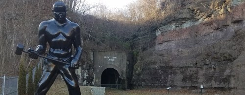 A large black statue of a muscular man with a hammer stands near the entrance of a tunnel in a rocky landscape.