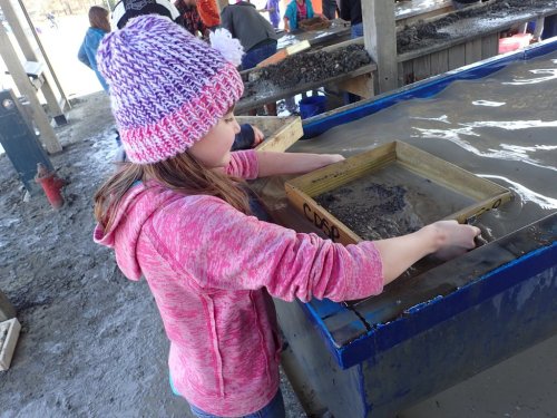 A girl in a pink sweater and knitted hat sifts through water and mud at a table, searching for treasures.