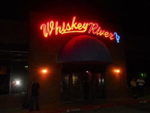 A brick building at night with a bright neon sign reading "Whiskey River" and a person standing outside.