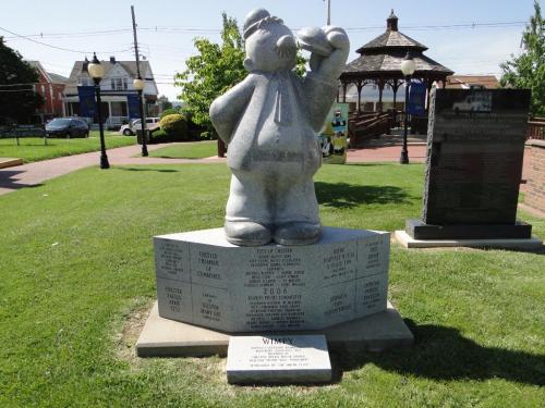 A stone statue of a bear holding a hat, surrounded by plaques and greenery in a park setting.