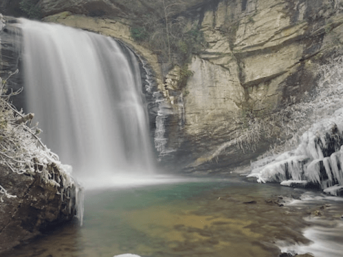 A serene waterfall cascades over rocky cliffs into a tranquil pool, surrounded by icy formations and lush greenery.